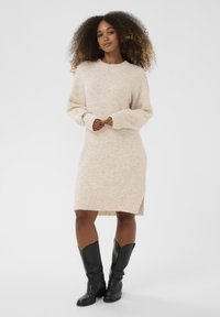 Bež pleteni džemper haljina dugih rukava, s rebrastim rubom i bočnim prorezima. U kombinaciji s crnim čizmama do koljena. Mekana tekstura, opušteni kroj.