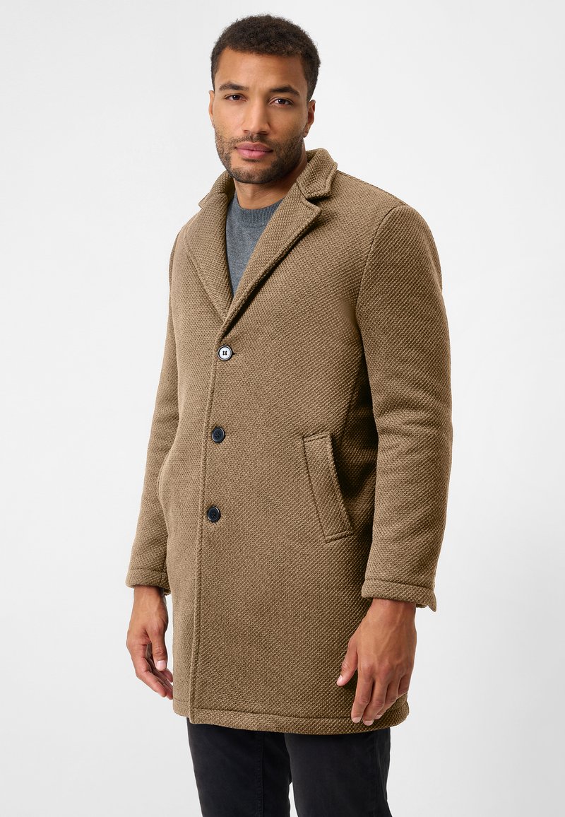 Brun tekstureret uldfrakke med sjalskrave, tre sorte knapper og sidelommer, båret over en grå sweater og sorte bukser.