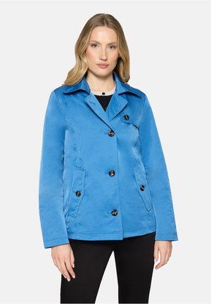 Kobieta ubrana w jaskrawo niebieską kurtkę zapinaną na guziki z kołnierzem i przednimi kieszeniami, stojąca na tle jednolitego jasnego tła.