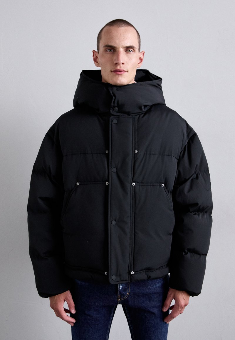 Mann in schwarzer Steppjacke mit hohem Kragen und blauen Jeans, der vor einem schlichten hellgrauen Hintergrund steht.