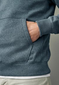 Sudadera gris con un bolsillo tipo canguro, puños y dobladillo de canalé. La tela parece suave con una textura lisa, contrastando con una camiseta más clara debajo.