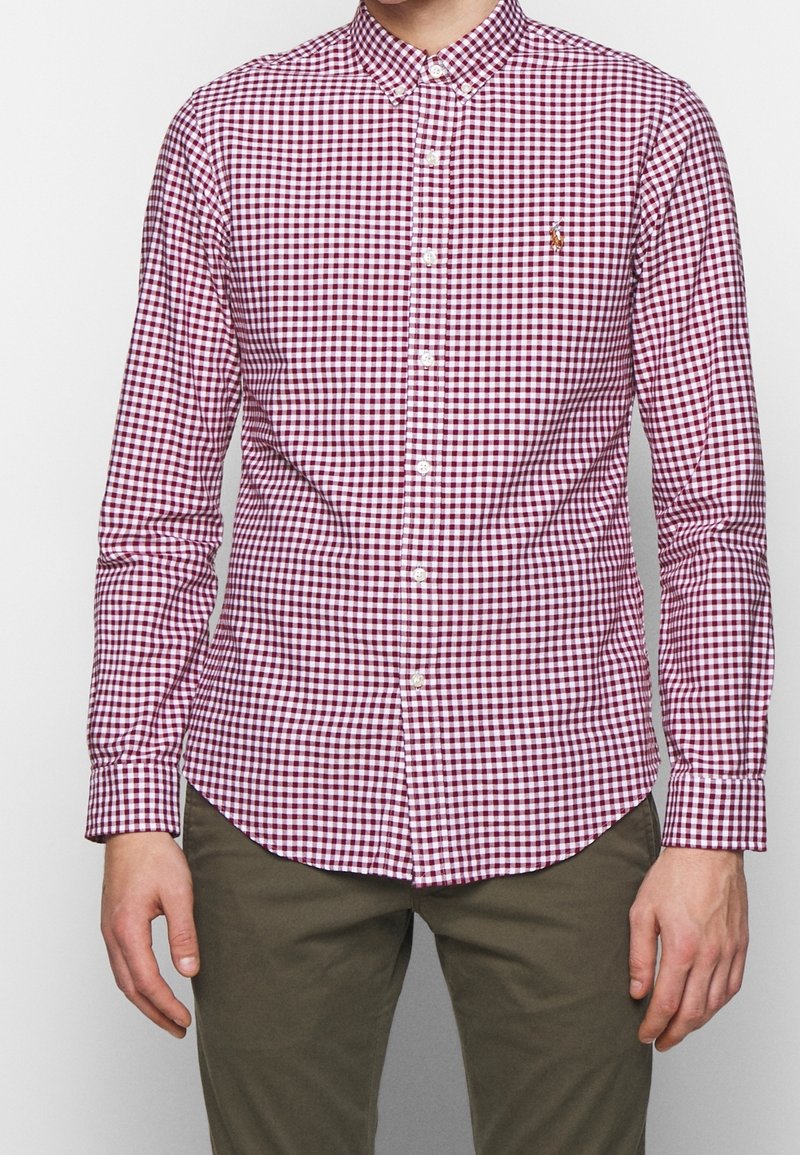 Camisa de manga larga con botones en patrón de cuadros vichy rojo y blanco, que presenta un cuello clásico y cierre frontal con botones, fabricada con una tela de textura suave.