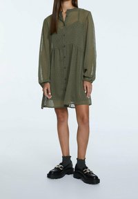 Khakigrünes Hemdkleid mit Knopfleiste, langen transparenten Ärmeln, strukturiertem Stoff und gerafftem Taillenbereich. Kombiniert mit schwarzen Schuhen und grauen Socken.