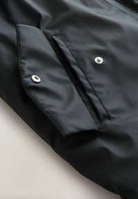 Juodas neperšlampamas švarkas su lygiu paviršiumi, slepiamu užtrauktuku, spaudžiama sagtele ir šonine kišene su plokščiu dizainu bei metaliniais akcentais.