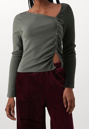 Woman wearing an olive green long-sleeve top with side buttons and burgundy velvet pants, standing against a plain background.