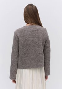 Ruskea, teksturoitu neulepusero, jossa on pitkät hihat ja pyöreä pääntie, lyhyt malli, joka on pukeutunut ilmavan valkoisen mekon päälle.