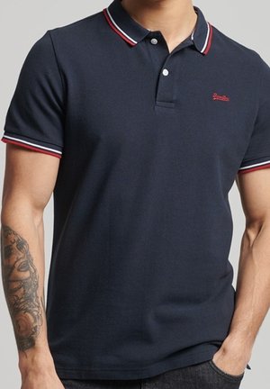 Mand iført en marineblå polo med krave og ærmekanter i røde, hvide og marineblå striber samt et rødt broderet logo på brystet.
