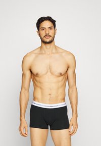 Schwarze Baumwoll-Boxerbriefs mit einem weißen Bund, auf dem das Logo "TOMMY HILFIGER" zu sehen ist. Das Modell steht frontal vor einem neutralen grauen Hintergrund.