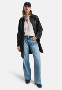 Cappotto di pelle nera sopra una camicia a righe rosa con bottoni, abbinato a jeans a gamba larga blu chiaro e scarpe navy, su uno sfondo bianco.