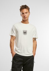 Vit bomull t-shirt med en svart Bundeswehr-örns logo i mitten. Korta ärmar och rund halsringning fulländar designen.