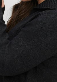Maglione nero lavorato a maglia con una finitura testurizzata, caratterizzato da un colletto ripiegato e maniche lunghe. Il tessuto ha una leggera lucentezza.