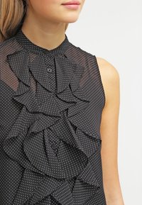 Mujer con blusa sin mangas negra con lunares blancos y detalles de volantes en capas en la parte delantera.