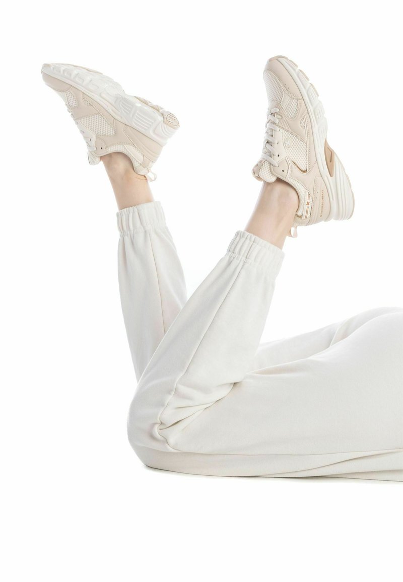 Zapatillas deportivas beige con detalles de malla y cuero, que presentan una gruesa suela blanca. Combinadas con pantalones de chándal de color crema con puños elásticos.