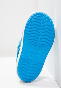 Sininen kengän ulkopohja, jossa on siksak-kuvio, valkoisella reunalla ja keltaisella korostuksella. Materiaali näyttää kumimaiselta, suunniteltu pitävyyttä varten.