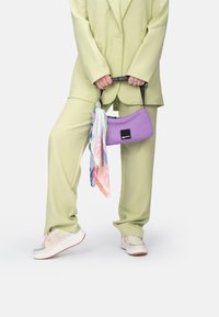 Sac à main violet au design épuré, associé à un foulard pastel, porté avec un costume vert clair et des baskets épaisses avec une semelle texturée.