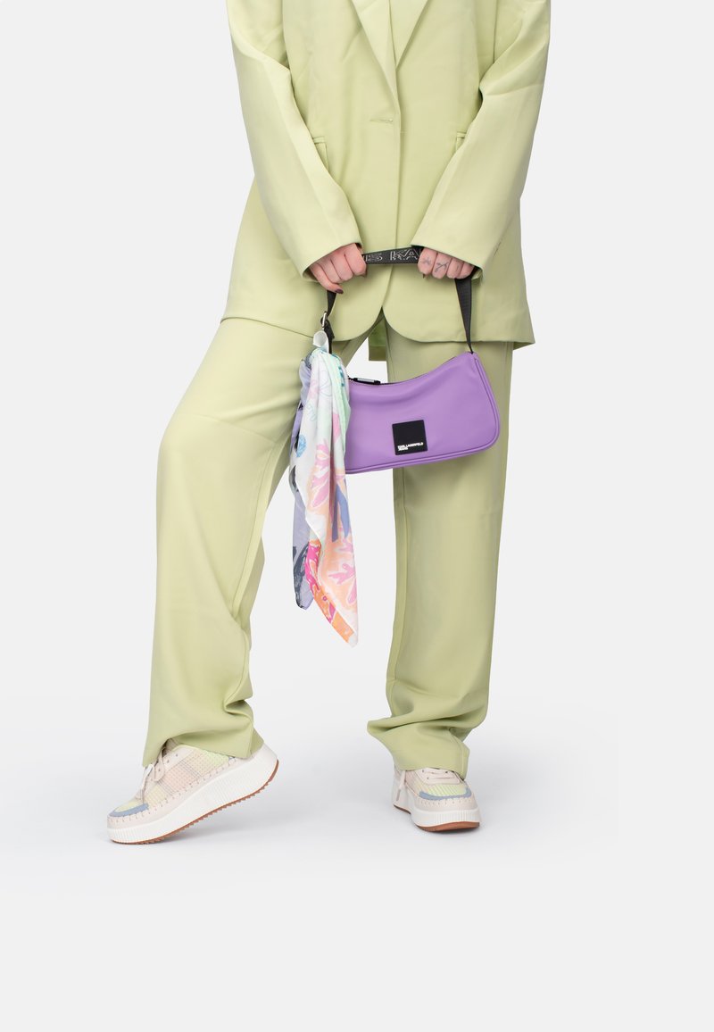 Sac à main violet au design épuré, associé à un foulard pastel, porté avec un costume vert clair et des baskets épaisses avec une semelle texturée.