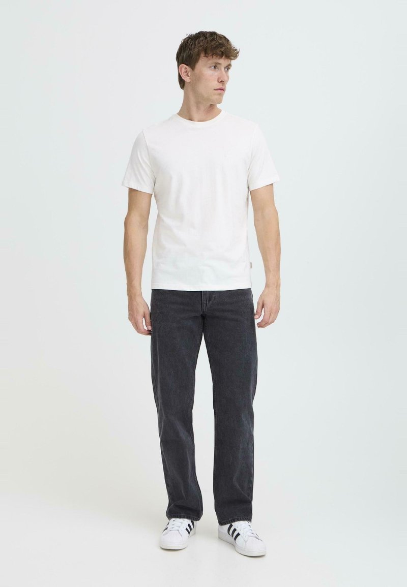 Camiseta blanca de manga corta combinada con jeans gris oscuro. El modelo está de pie sobre un fondo sencillo, mostrando un atuendo casual con zapatillas blancas.