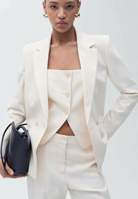 Witte op maat gemaakte blazer en cropped top met drie knopen; gecombineerd met bijpassende broek. Model houdt een zwarte gestructureerde handtas vast.