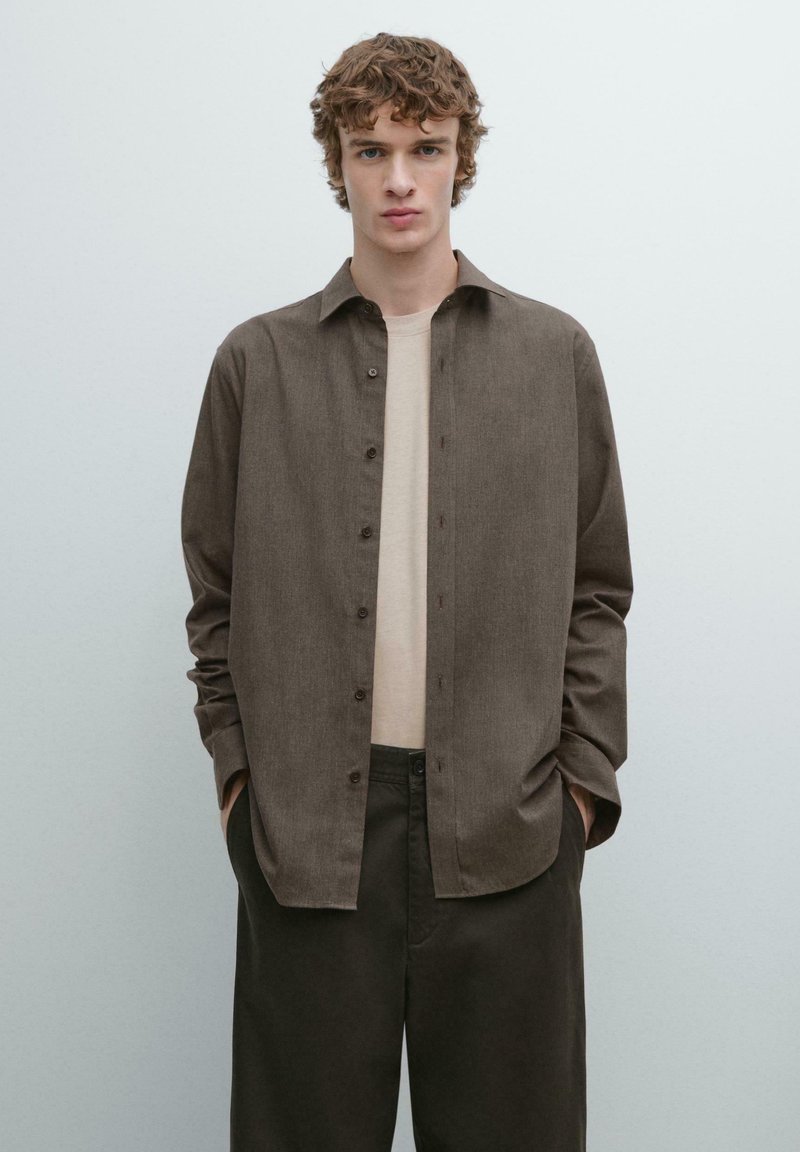 Brun skjorte med knapper, løs pasform og lange ærmer, lavet af struktureret stof, kombineret med en lysefarvet T-shirt og mørke bukser.