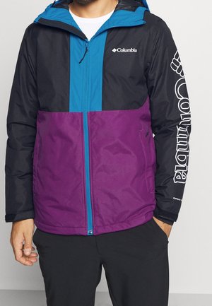 Homme portant une veste Columbia noire, bleue et violette avec un logo sur la poitrine et un grand texte sur la manche droite, debout les mains près des poches.