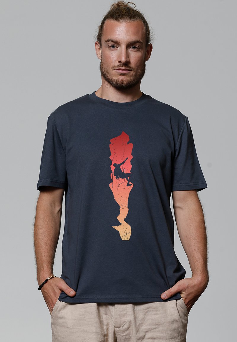 Homme aux cheveux attachés portant un T-shirt bleu foncé affichant la silhouette d'un grimpeur sur une formation rocheuse verticale stylisée allant de l'orange au rouge.