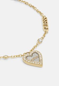 Gold-tone necklace featuring a heart-shaped pendant with a crystal embellishment, intricate design, and the word "GUESS." Chain link design.