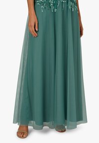 Długa sukienka w kolorze morskim z cekinami w talii, z przezroczystą nakładką na spódnicy, noszona z odkrytymi beżowymi sandałami.