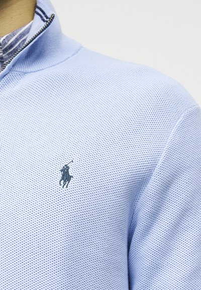 Ανοιχτό μπλε υφασμάτινο πουλόβερ με φερμουάρ έως το στήθος, με κεντημένο λογότυπο παίκτη πόλο σε σκούρο μπλε χρώμα στη αριστερή πλευρά του στήθους, φορεμένο πάνω από ριγέ πουκάμισο με γιακά.