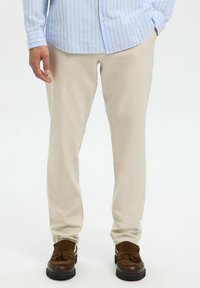 Pantalones beige a medida hechos de una tela suave, con un corte slim. Llevados con una camisa de botones a rayas de color azul claro y mocasines marrones.