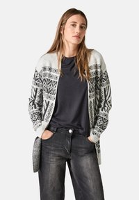 Grå och svart mönstrad kofta med långa ärmar, öppen front och en texturerad finish, bärs över en svart skjorta och mörka jeans.
