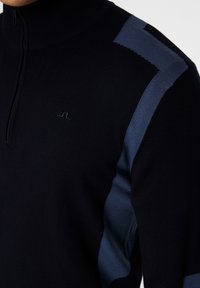 Marinblått stickat tröja med halvdragkedja i kragen, med blå detaljer på axlarna och en liten logotyp på bröstet. Slät textur.