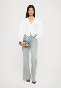 Witte gestructureerde cropped jas met pofmouwen, gecombineerd met lichtblauwe flared jeans. De houder heeft een blauw en oranje ontwerp.