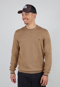 Pull en tricot marron clair avec des manches longues, un col rond, des accents foncés aux poignets et à l'ourlet, arborant un petit logo sur la poitrine.