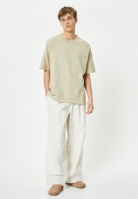 Beige ylisuurennettu puuvillateeppari, jossa on pyöreä kaula-aukko ja lyhyet raglan-hihat, yhdistettynä leveisiin valkoisiin housuihin ja vaaleanruskeisiin liukuhihnasandaaleihin.