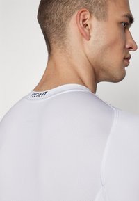 Hvid TECHFIT træningsshirt lavet af tekstureret materiale, med rund halsudskæring og glatte sømme, samt logo placeret på ryggen.
