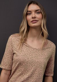 Nuori nainen, jolla on pitkät vaaleat hiukset, pukeutuneena vaaleanpunaiseen ja ruskeaan kuvioituun lyhythihaiseen toppiin tummanharmaata taustaa vasten.