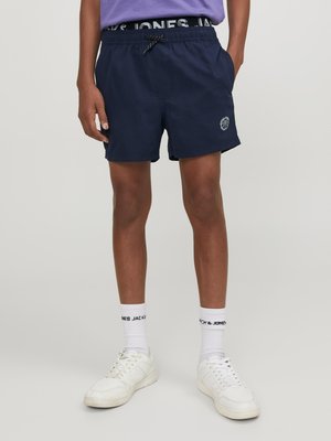 Marineblauwe zwembroek met een elastische tailleband, verstelbare koord, zijzakken en een logo-opdruk op de zoom. Witte sneakers en sokken.
