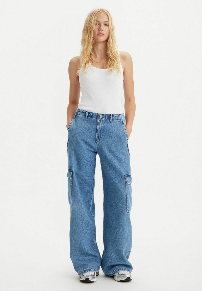 Hellblaues Denim-Cargo-Hose mit weitem Bein, Seitentaschen und leicht ausgefransten Säumen. Kombiniert mit einem enganliegenden weißen Tanktop.