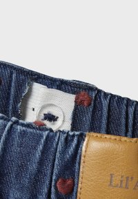 Denim fabric with gathering, featuring small red heart embroidery and a tan leather patch with the text "Lil’ A." Secure button closure visible.