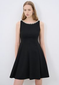 Vestido negro sin mangas con una falda acampanada, decorado con un escote de cuentas y una textura de tela suave. Silueta simple y ajustada.