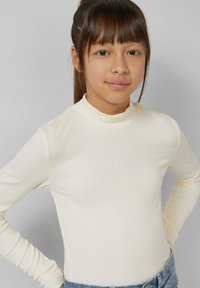 Kremowa, prążkowana koszulka z długim rękawem i wysokim dekoltem. Materiał jest gładki i dopasowany, zestawiona z niebieskimi jeansowymi spodniami.