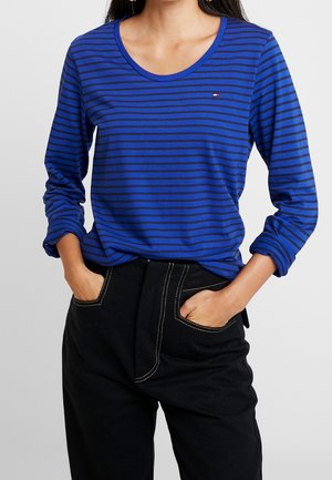 Kobieta w niebieskiej koszuli z długim rękawem z cienkimi czarnymi poziomymi paskami oraz czarnych spodniach, trzymająca ręce w kieszeniach.