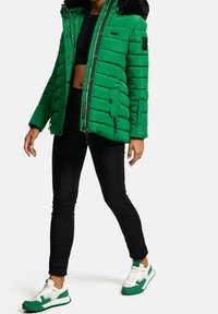 Chaqueta acolchada verde con capucha forrada de piel, cierres de cremallera y botón, puños acanalados y un parche negro en la manga. Lleva puesta sobre pantalones y zapatos negros.