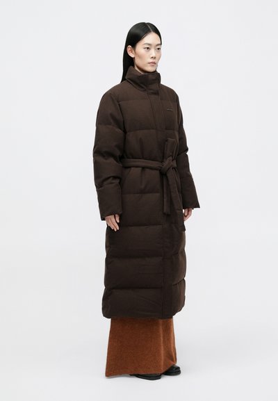 Brun polstret lang frakke med høj krave, belagt talje og vandrette stikninger, parret med en lang brun nederdel og sorte støvler.