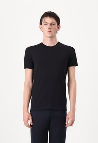 Juodas trumpas rankovės marškinėlis, pagamintas iš lygaus audinio, su apvaliu kaklu ir prigludusia forma, derinamas su tamsiomis kelnėmis prieš baltą foną.