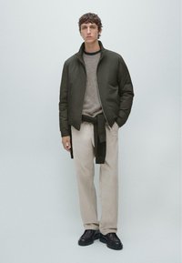 Giacca bomber leggera verde scuro sopra un maglione lavorato a maglia color cammello, abbinata a pantaloni beige chiaro e scarpe nere, design minimalista.