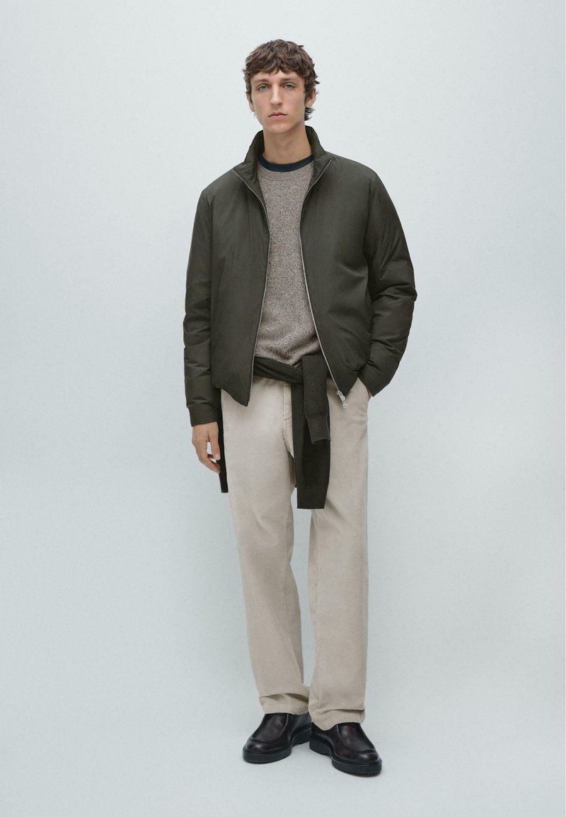 Giacca bomber leggera verde scuro sopra un maglione lavorato a maglia color cammello, abbinata a pantaloni beige chiaro e scarpe nere, design minimalista.