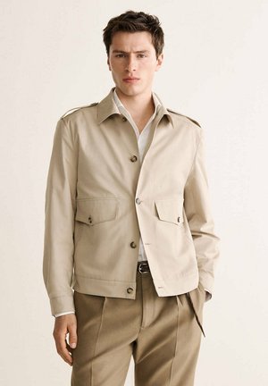 Junger Mann trägt eine beige Knopfleiste Jacke mit zwei Vorder-Taschen über weißem Hemd und brauner Hose, steht vor einfarbigem Hintergrund.