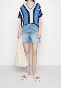 Haut en tricot à col en V rayé bleu, crème et marine, associé à un short en denim bleu clair et des sandales à plateforme blanches, tenant un sac fourre-tout crème.