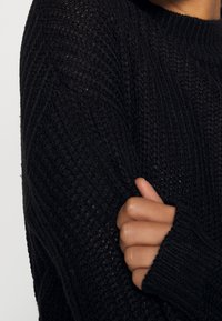 Personne croisant les bras, portant un pull en maille noire texturée avec des manches longues et des poignets côtelés.